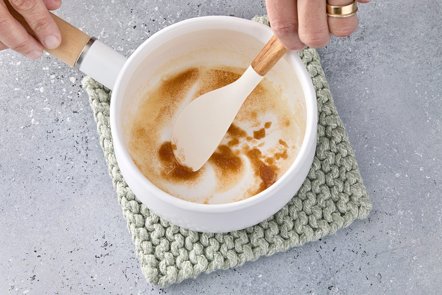 A person stirs browned butter in a white saucepan using a spatula, placed on a knitted green potholder. The background is a textured gray surface.