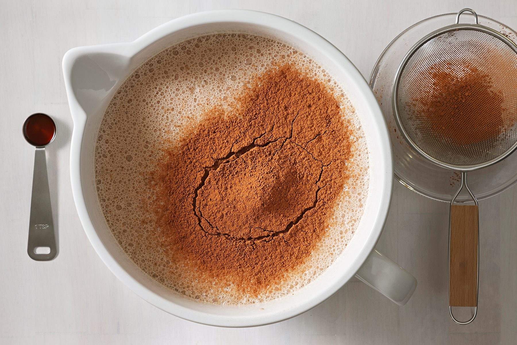 A mixing bowl filled with liquid and a heap of cocoa powder on top, next to a teaspoon of vanilla extract and a fine mesh sieve with cocoa residue on a light gray surface.