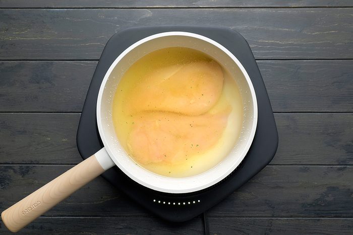 overhead shot of a white pan with a wooden handle on a black induction cooktop; the pan is filled with broth and two chicken breasts; the cooktop is on a black wooden surface