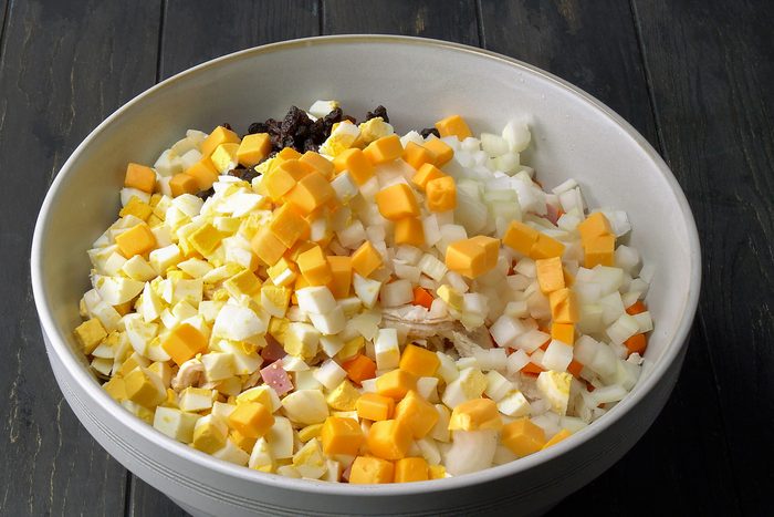 3/4th shot of a white bowl filled with ingredients for a salad, likely chicken salad; the bowl sits on a dark wood surface
