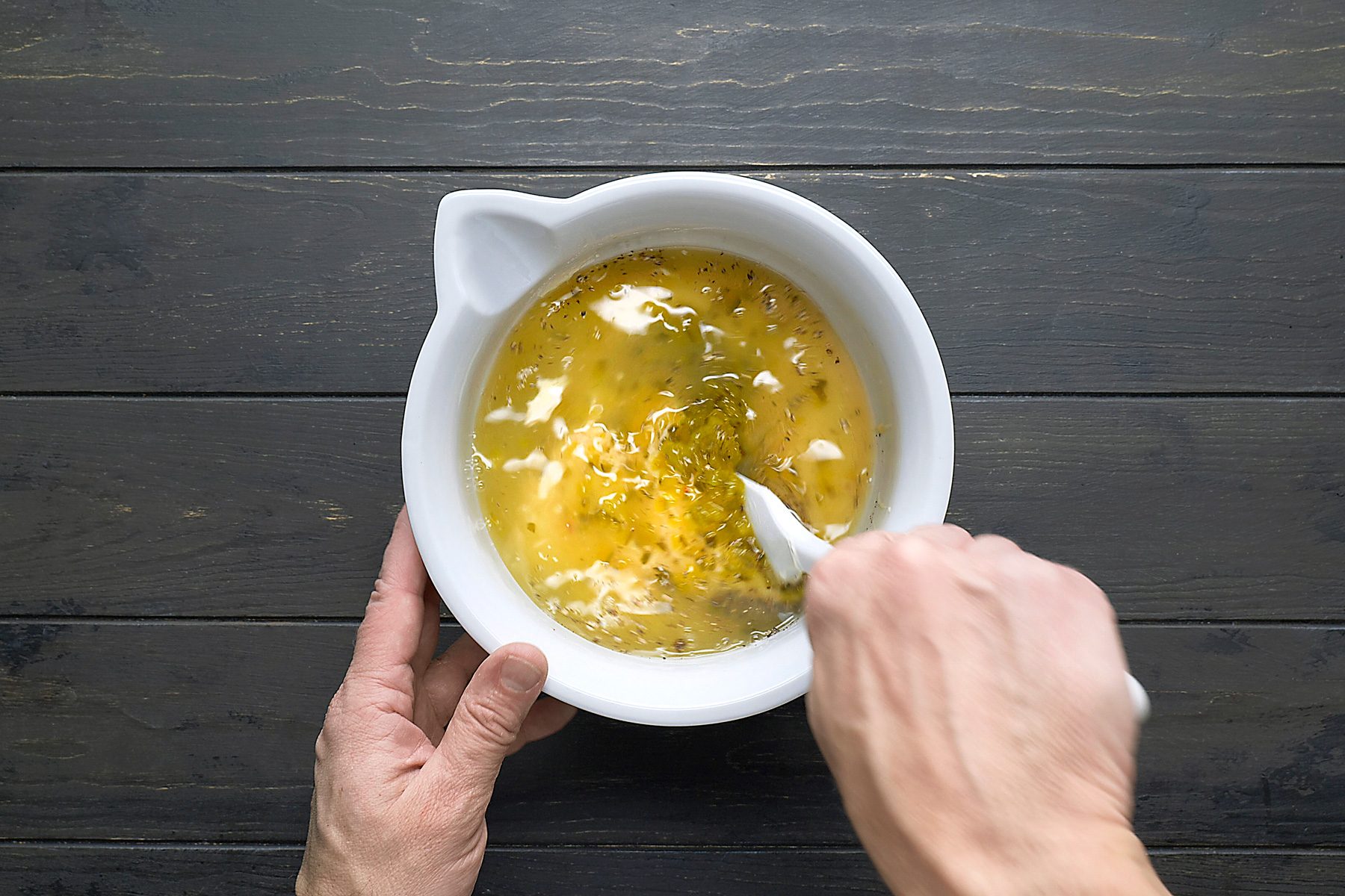 overhead shot of a white bowl filled with a yellow liquid and green flecks; a person is holding the bowl with their left hand and stirring the liquid with a white spoon in their right hand; the background is a dark wooden surface.