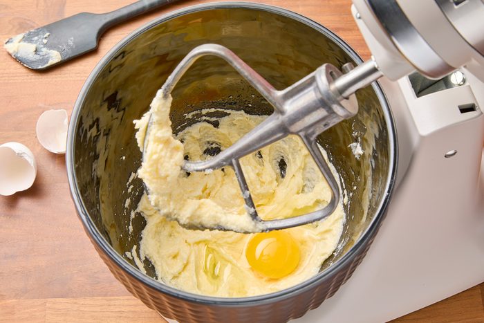 Overhead shot of a stand mixer with a paddle attachment mixing a bowl of batter. The batter contains a single egg yolk in the center, surrounded by white batter, with a bit of batter stuck to the paddle. A spatula is positioned to the left of the bowl, and a wooden table is visible beneath the mixer and spatula. Partially visible eggshells are in the bottom left corner of the image.