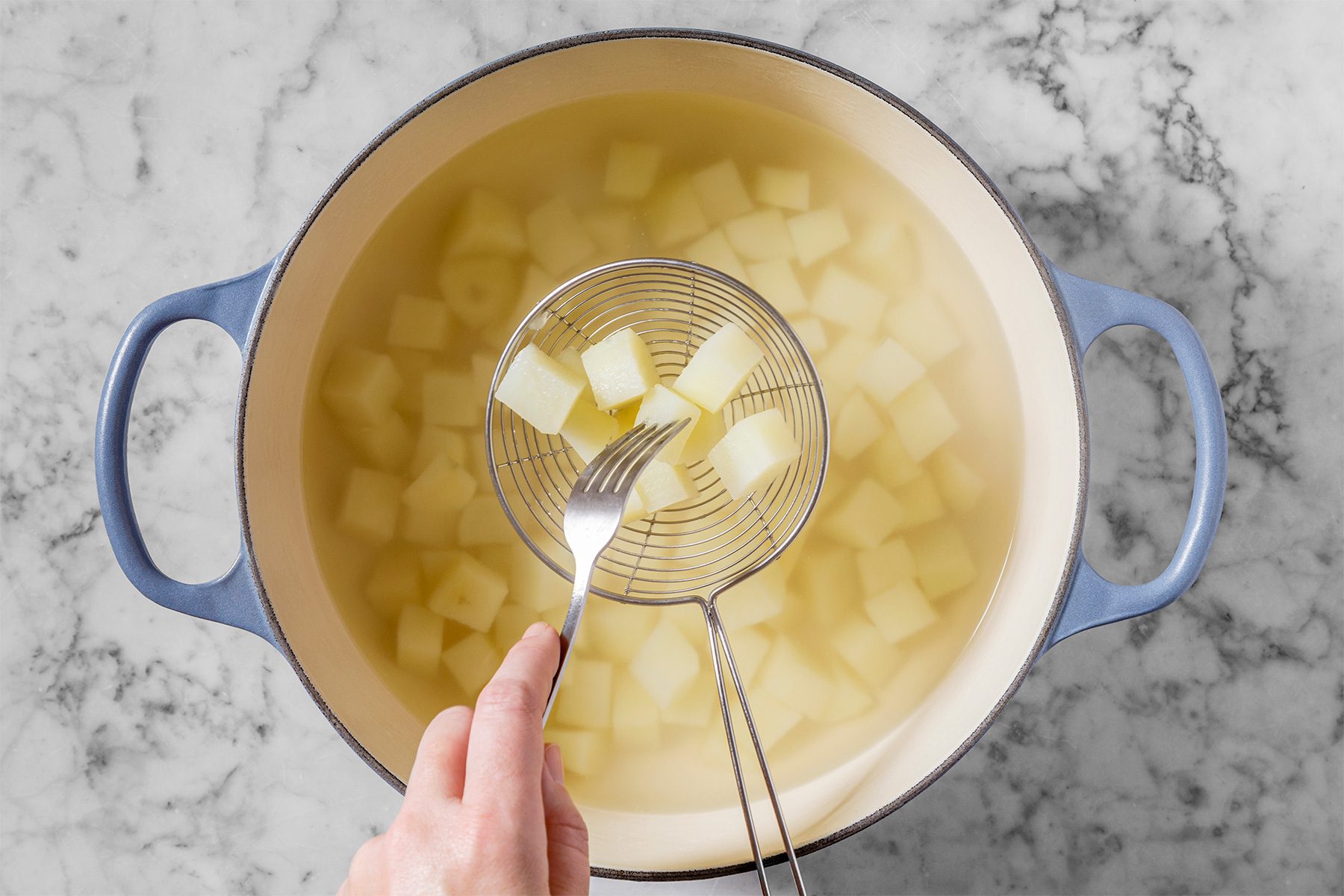A hand holds a slotted spoon lifting diced potato pieces from a pot of water. The pot is large, round, and has blue handles, placed on a marble countertop.