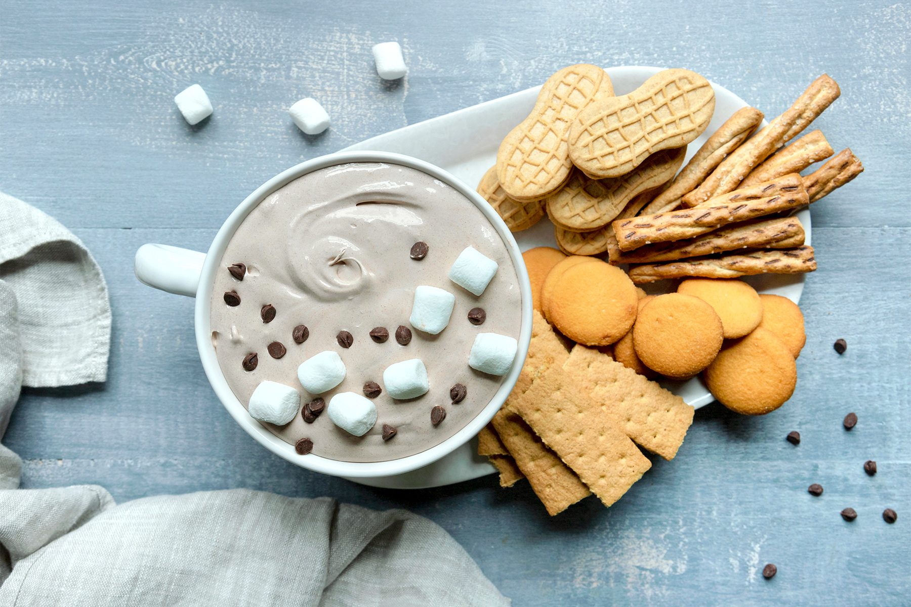 A bowl of creamy dip topped with marshmallows and chocolate chips, surrounded by various snacks including peanut-shaped cookies, breadsticks, round cookies, and graham crackers on a light blue textured surface.