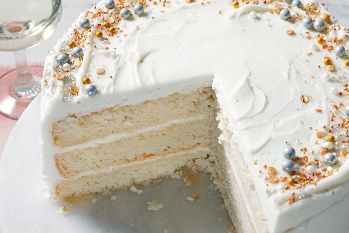 A three-layer vanilla cake with white frosting on a marble surface. The top is decorated with silver balls and golden sprinkles. A slice is missing from the cake. A glass of white wine is partially visible in the background.