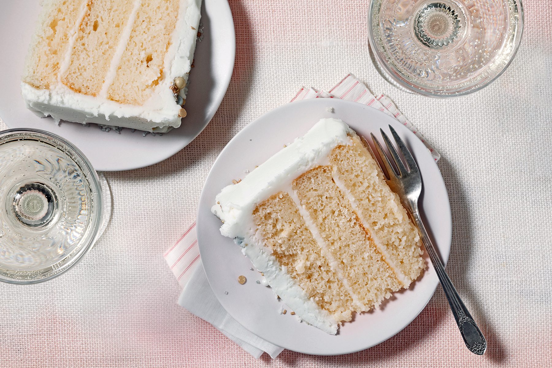 A slice of vanilla layer cake with white frosting sits on a white plate beside a fork. Another slice is partially visible on a separate plate. Two clear glasses are nearby on a light fabric surface.