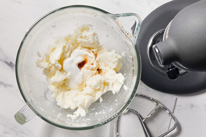 A glass mixing bowl with softened butter and powdered sugar being creamed together in a stand mixer. A splash of vanilla and a spoonful of yogurt are visible, with a mixing attachment resting nearby on a white marble countertop.