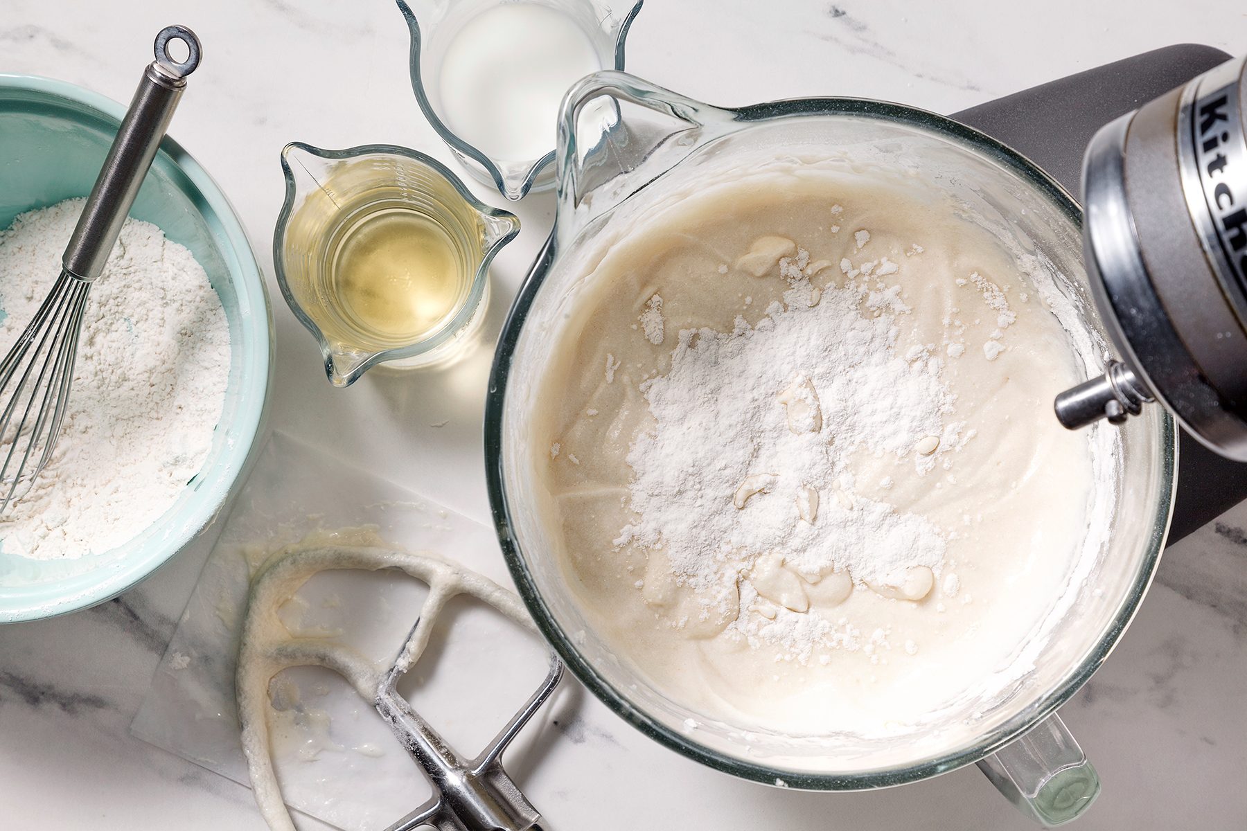A top view of a marble countertop with a mixing bowl on a stand mixer containing batter and flour. Nearby are a teal bowl with a whisk and flour, a measuring cup with milk, and one with oil. A beater attachment rests on the counter.