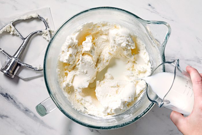 A hand pours milk into a glass mixing bowl filled with creamy frosting. A metal beater rests on a nearby surface. The bowl sits on a marble countertop.