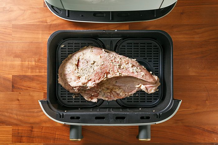 A seasoned raw piece of meat is placed inside an air fryer basket on a wooden countertop, ready for cooking.