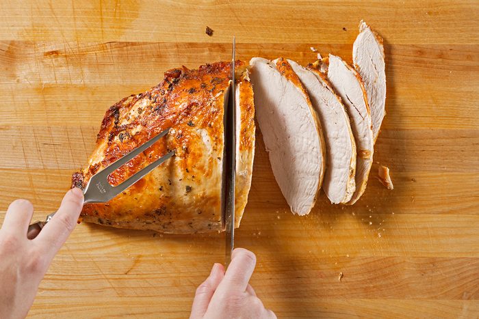 A person slices a cooked, golden-brown piece of turkey breast on a wooden cutting board, revealing several juicy, evenly cut slices. A fork secures the meat while a knife carves through it.