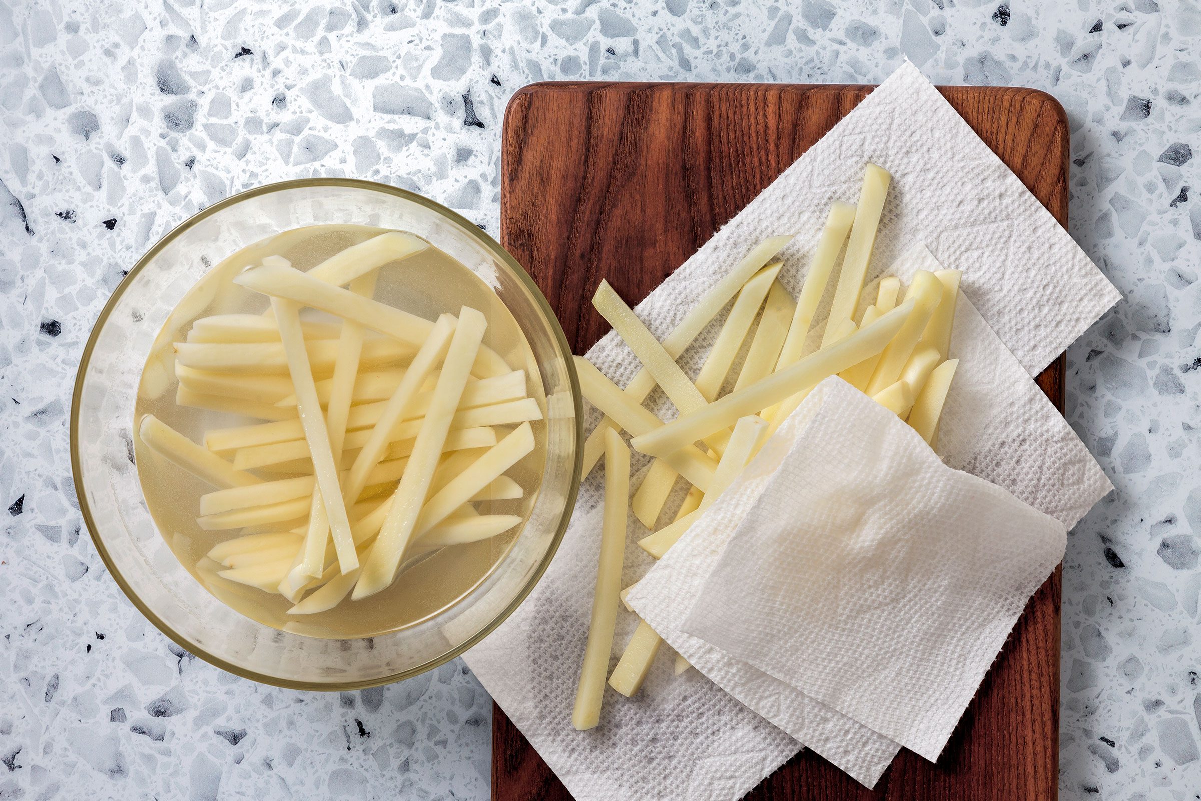 Potato strips covered with water in a bowl with potato strips being pat dried with kitchen napkin