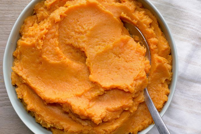 overhead shot of a bowl of mashed sweet potatoes, with a spoon resting on top; the sweet potatoes are a vibrant orange color, and the bowl is a pale blue-green;