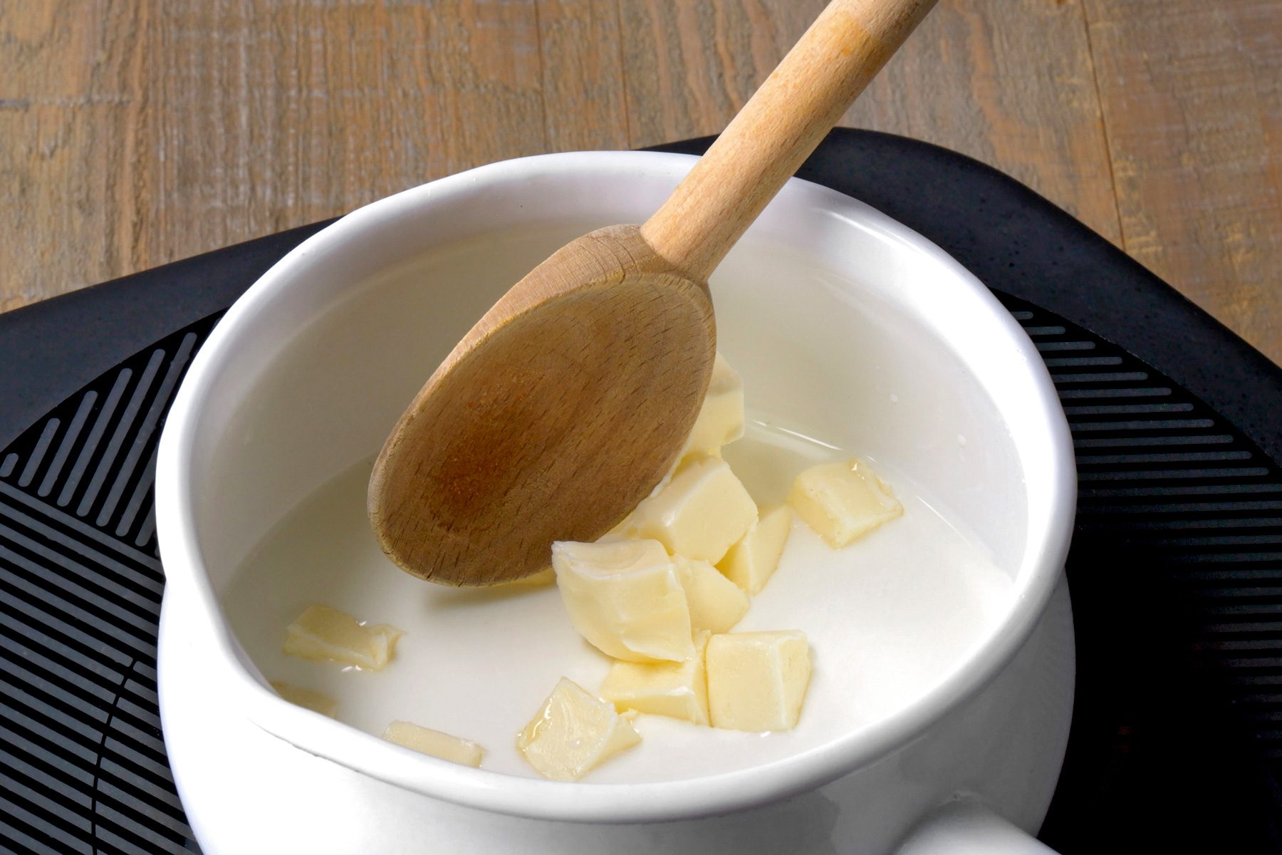 3/4th shot of a white ceramic bowl with a wooden spoon is on top of a black cooking mat, and a wooden table is in the background the bowl contains liquid and pieces of white butter; the spoon is sticking out of the bowl the spoon's handle is also made of wood; the background of the image is a wooden table