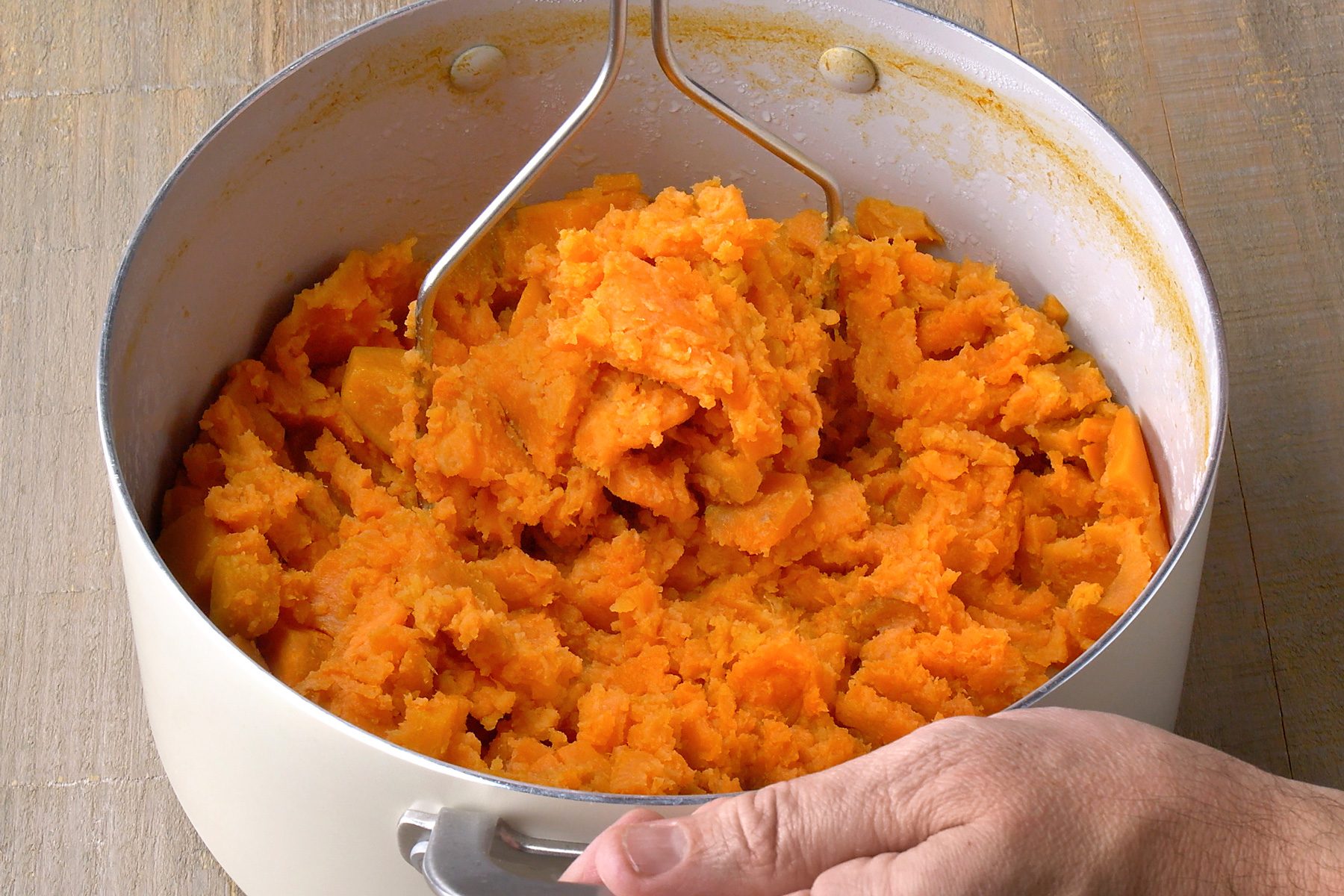3/4th shot of a pot containing mashed sweet potatoes, a hand is reaching into the pot to mix the potatoes, a potato masher is sitting inside the pot; The pot is made of white metal with a silver handle and is sitting on a light brown wooden surface