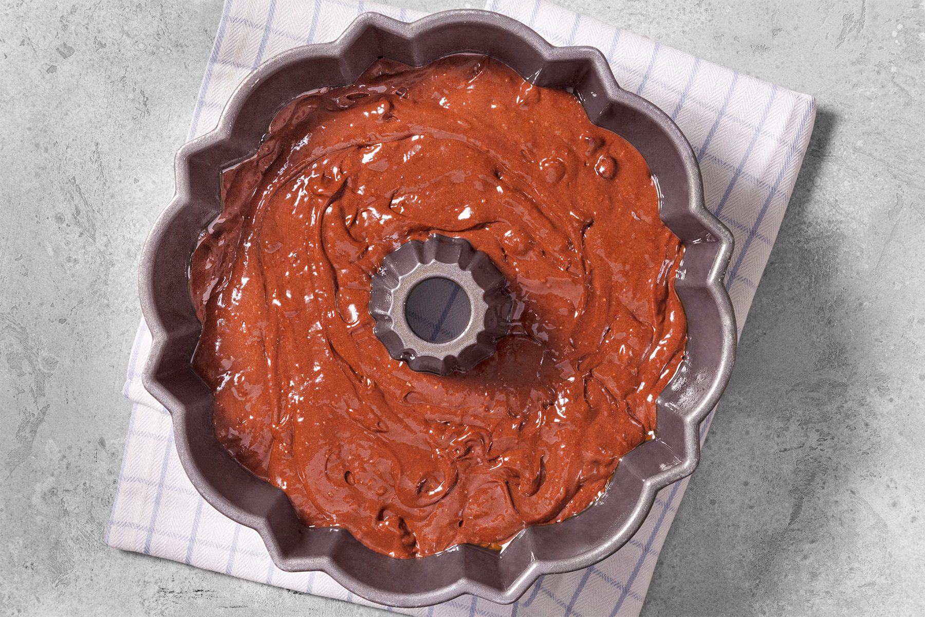 overhead shot of a chocolate cake batter in a bundt cake pan, ready to be baked; the pan is on a white and blue checkered kitchen towel, which is on a light gray countertop;
