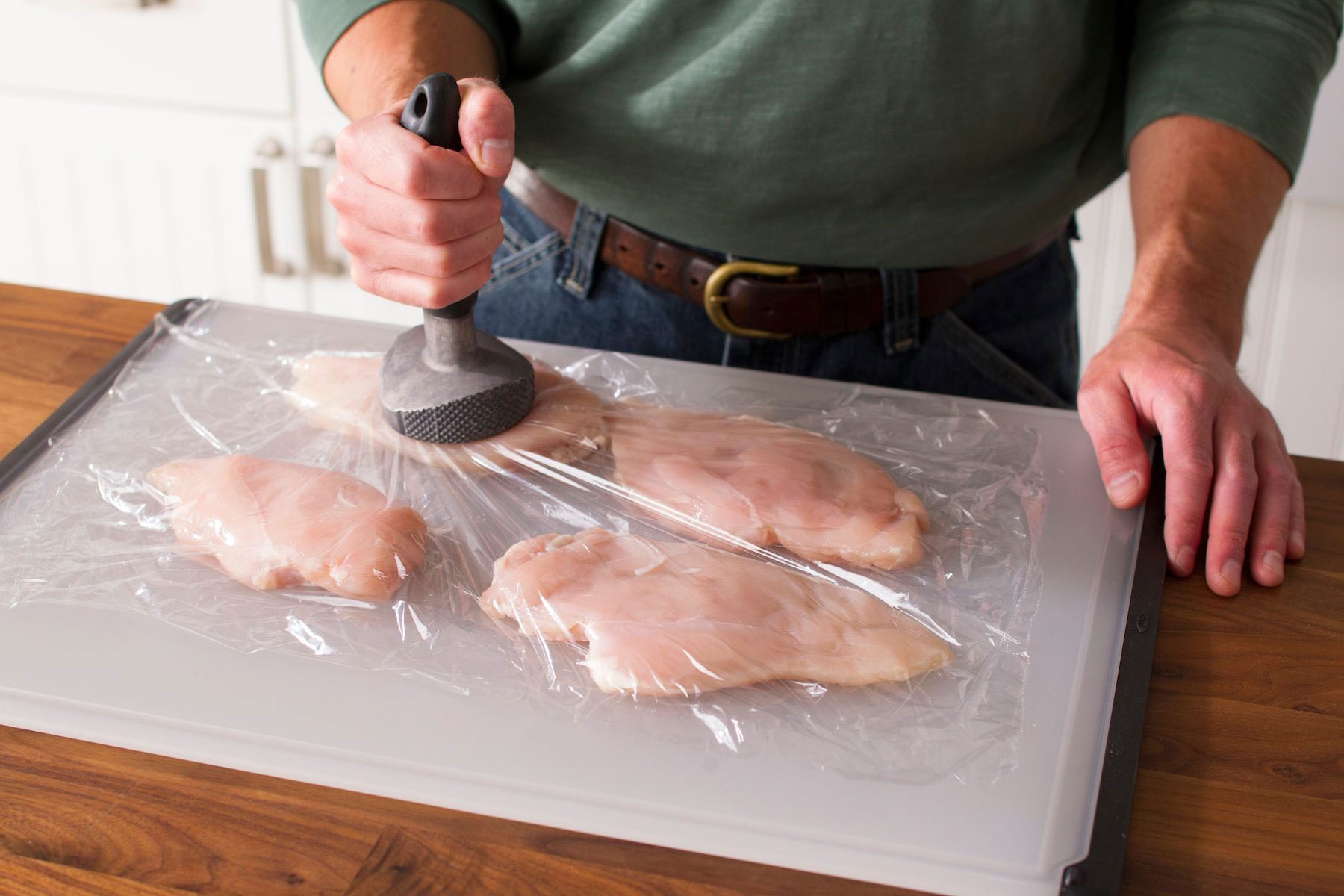 A person using a meat tenderizer to flatten raw chicken breasts on a cutting board, covered with plastic wrap. The countertop is wooden, and the individual is wearing a green top and jeans.
