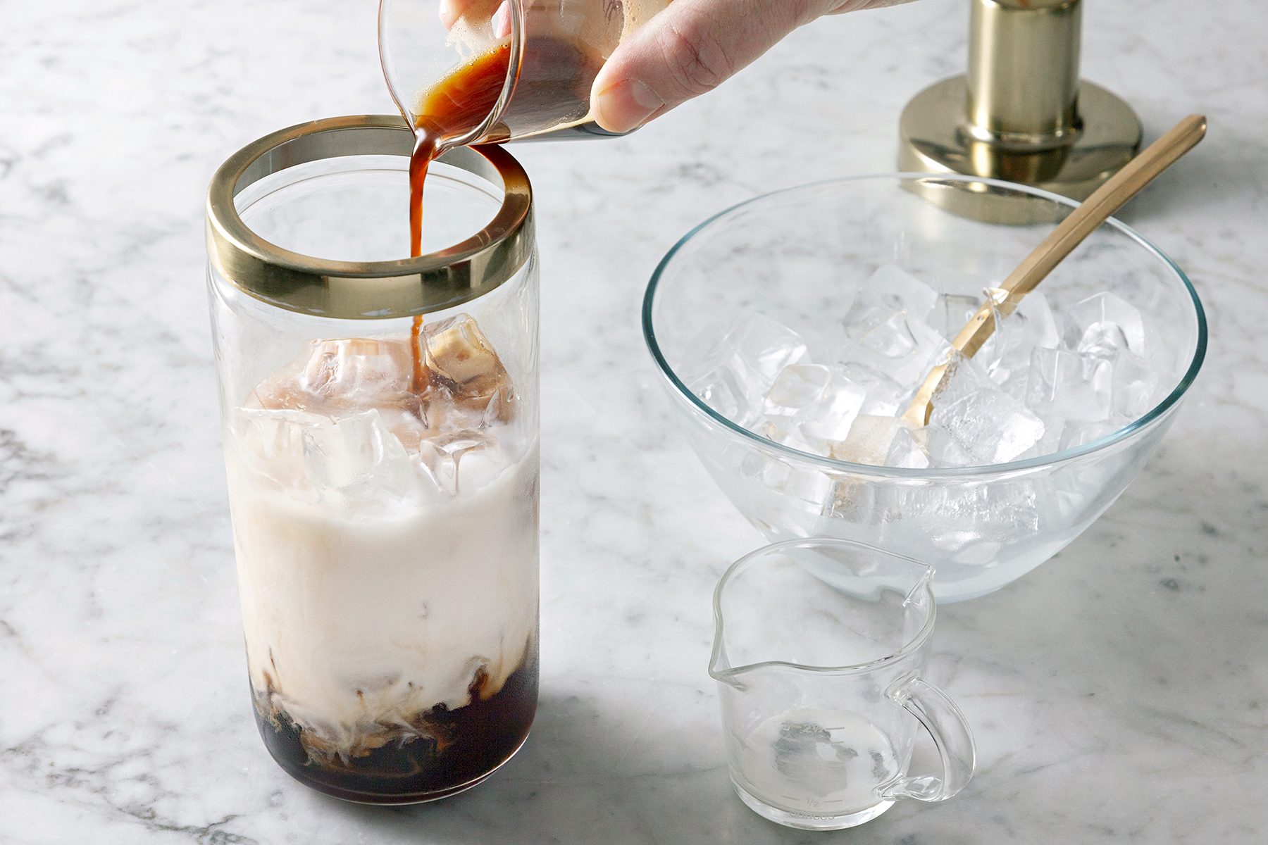 A hand pours espresso into a tall glass filled with milk and ice, set on a marble surface. Nearby, there's an empty glass bowl with ice cubes and a small glass pitcher.