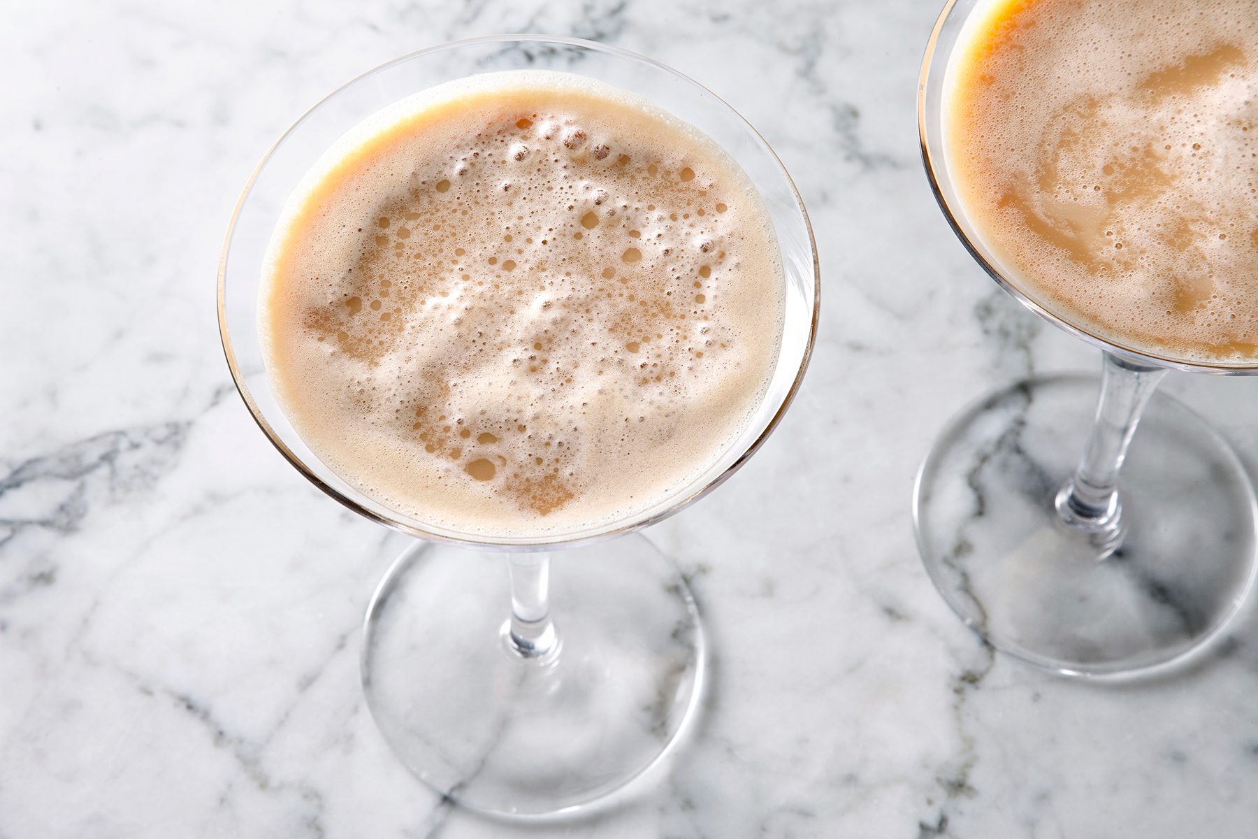 Two martini glasses filled with a frothy, light brown cocktail are placed on a white marble surface. The drinks have a creamy texture and are positioned side by side.