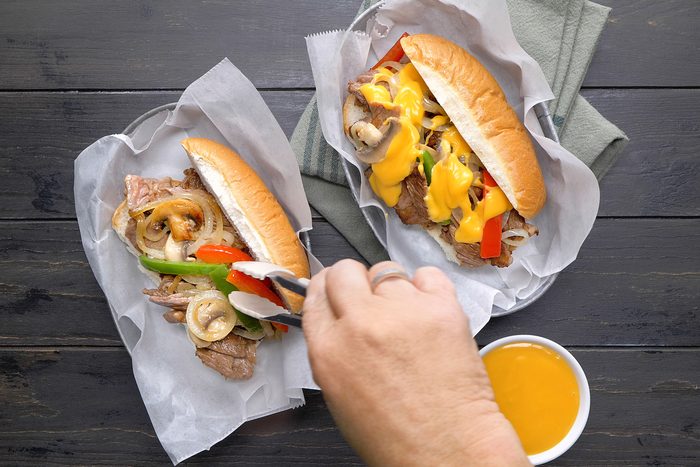 Assembling philly cheesesteak by adding ribeye steak in sliced roll topped with vegetables and cheese sauce