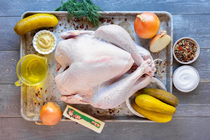 Raw turkey on a tray surrounded by ingredients: pickles, onions, a half lemon, dry spices, a dish of sugar, a bowl of mustard, a cup of olive oil, a stick of butter with a festive wrapper, and a sprig of dill.