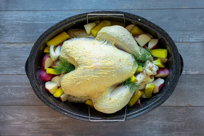 A seasoned raw turkey is placed in a roasting pan, surrounded by assorted chopped vegetables like potatoes and zucchini, garnished with fresh herbs, ready for cooking on a wooden surface.