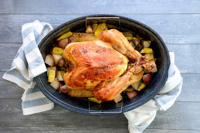A roasted chicken sits in a black roasting pan surrounded by roasted potatoes and sliced pickles. A striped kitchen towel is wrapped around the handles of the pan, which is placed on a gray wooden surface.