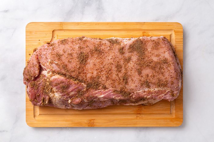 Rolled pork loin roast seasoned with a spice mixture on a wooden cutting board for step two of Pork Roast and Sauerkraut recipe for Taste of Home