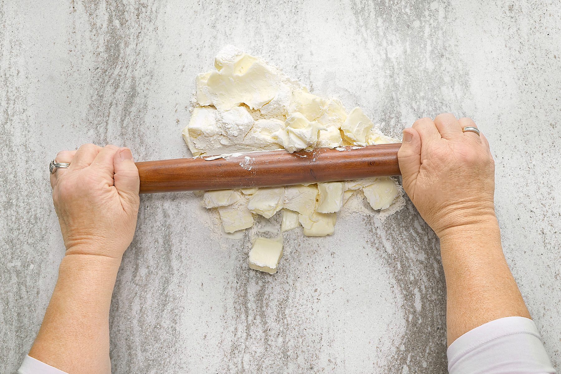 overhead shot of a person using a rolling pin on a countertop covered in flour and butter; the countertop is a light gray with a lot of speckles
