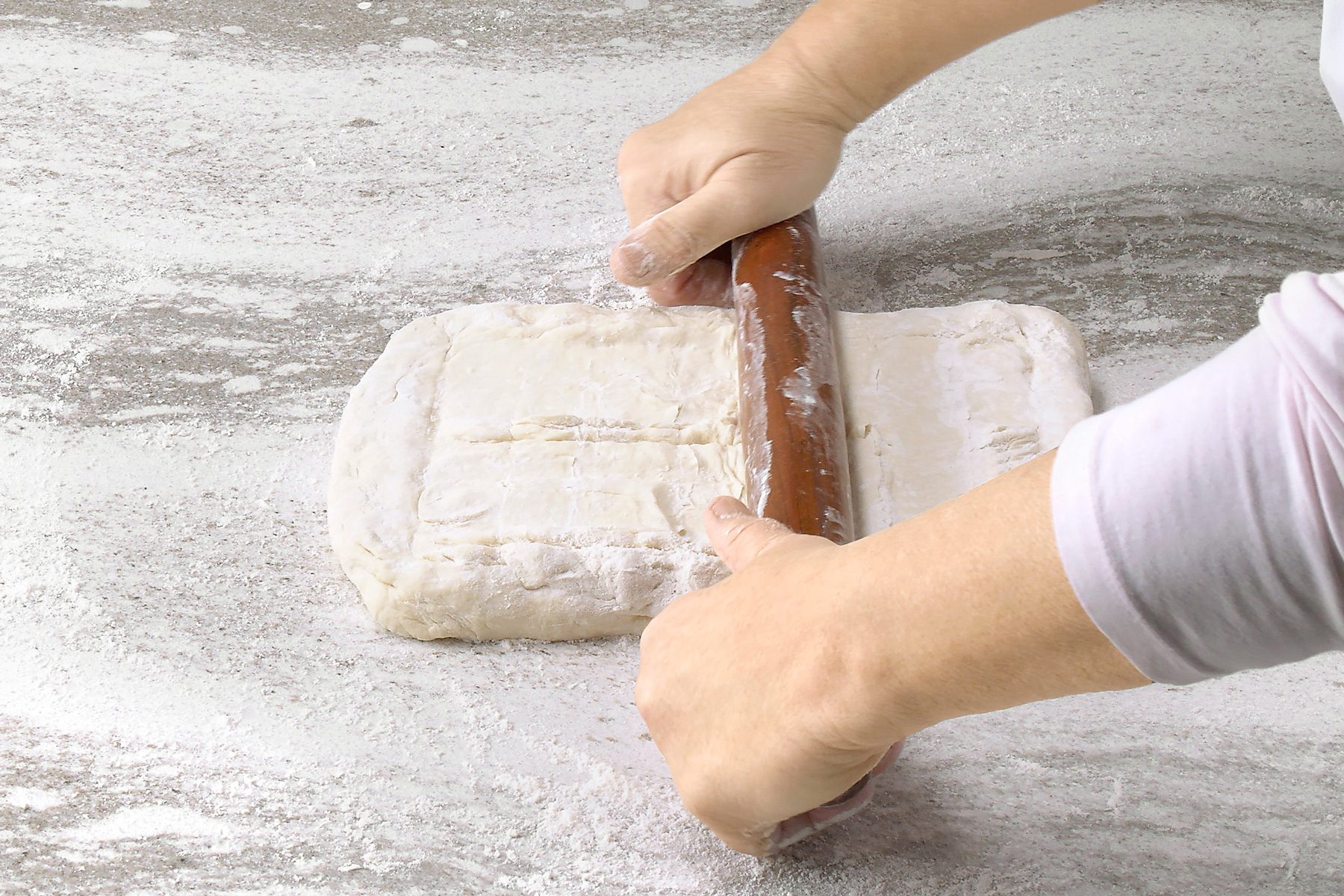 3/4th shot of a person is rolling out dough on a floured surface, using a wooden rolling pin; the dough is a rectangular shape