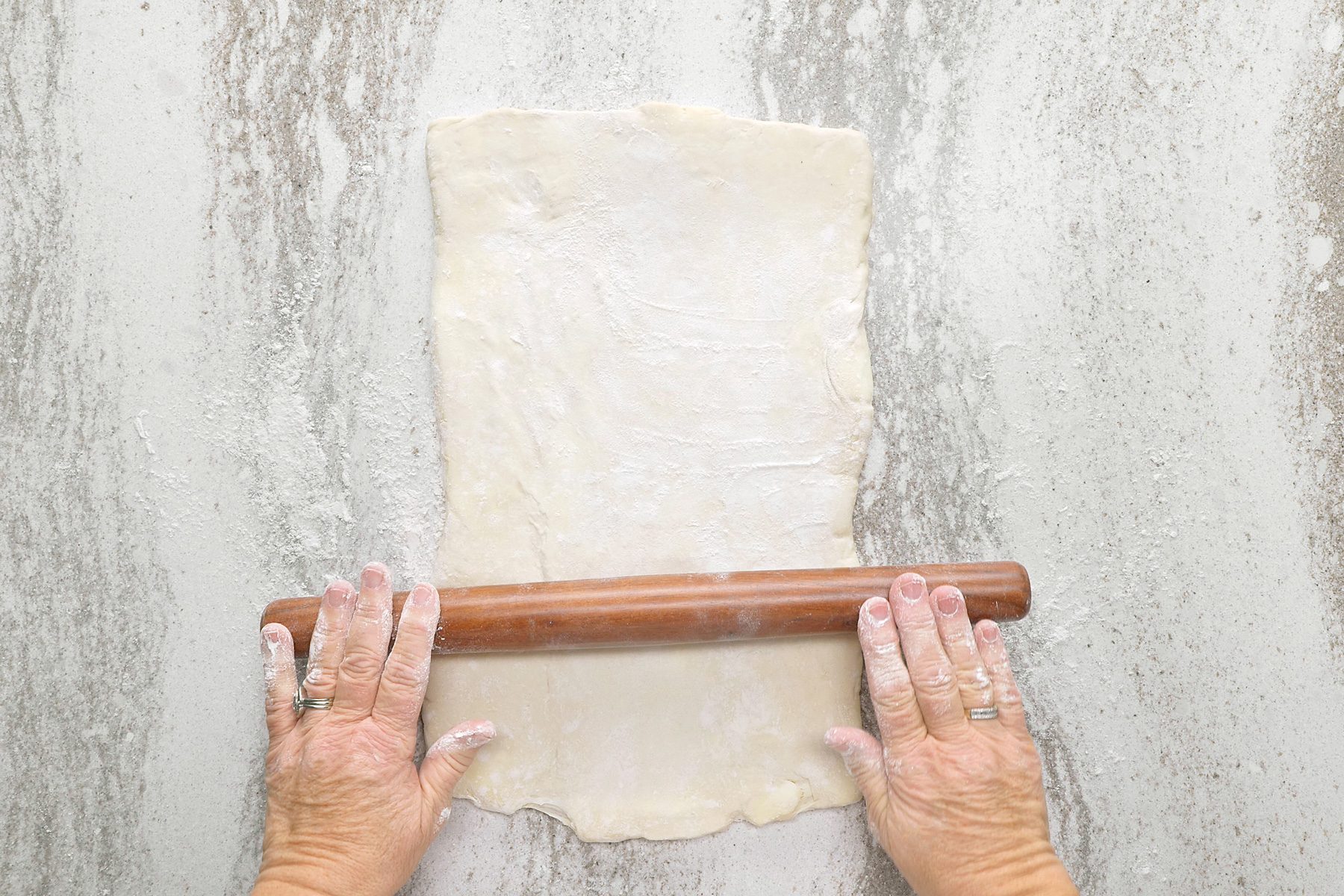 overhead shot of a person is rolling out dough on a floured surface, using a wooden rolling pin; the dough is a rectangular shape