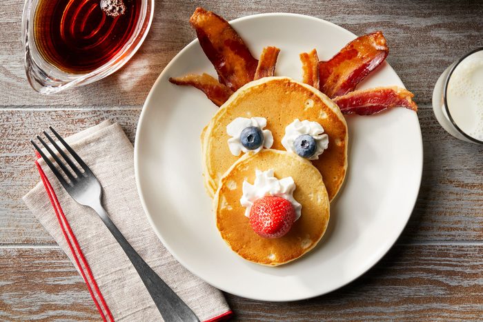 Assemble reindeer pancakes with whipped cream, blueberries, strawberries, and beacon