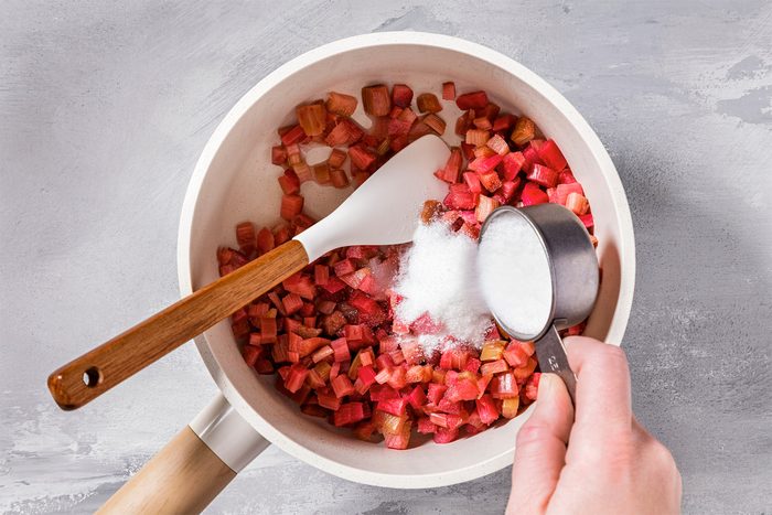 Overhead shot of a small saucepan; combine rhubarb; sugar and water; bring to a boil; spatula; grey surface;