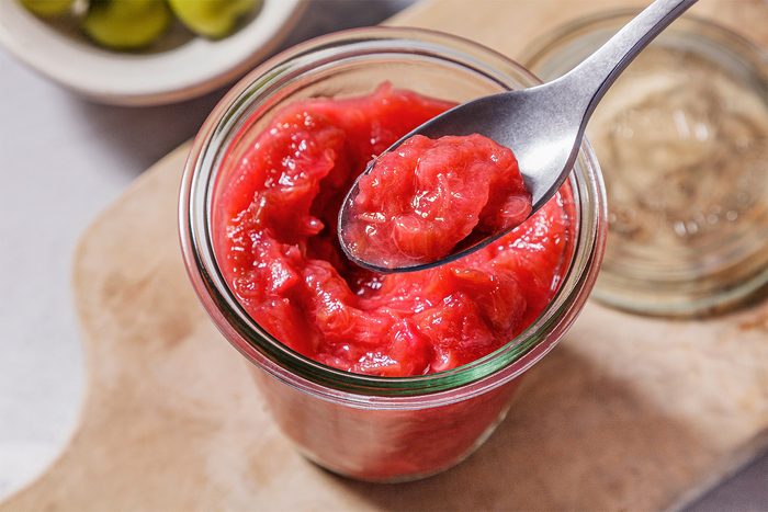 Overhead shot of Rhubarb Compote; in a glass jar; spoon; wooden tray; grey surface;