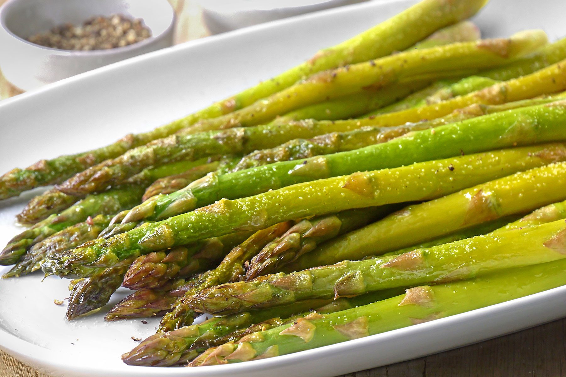 A platter of roasted asparagus is served on a white dish. The spears are drizzled with oil and lightly seasoned with pepper and salt. Small bowls of peppercorns are visible in the background.