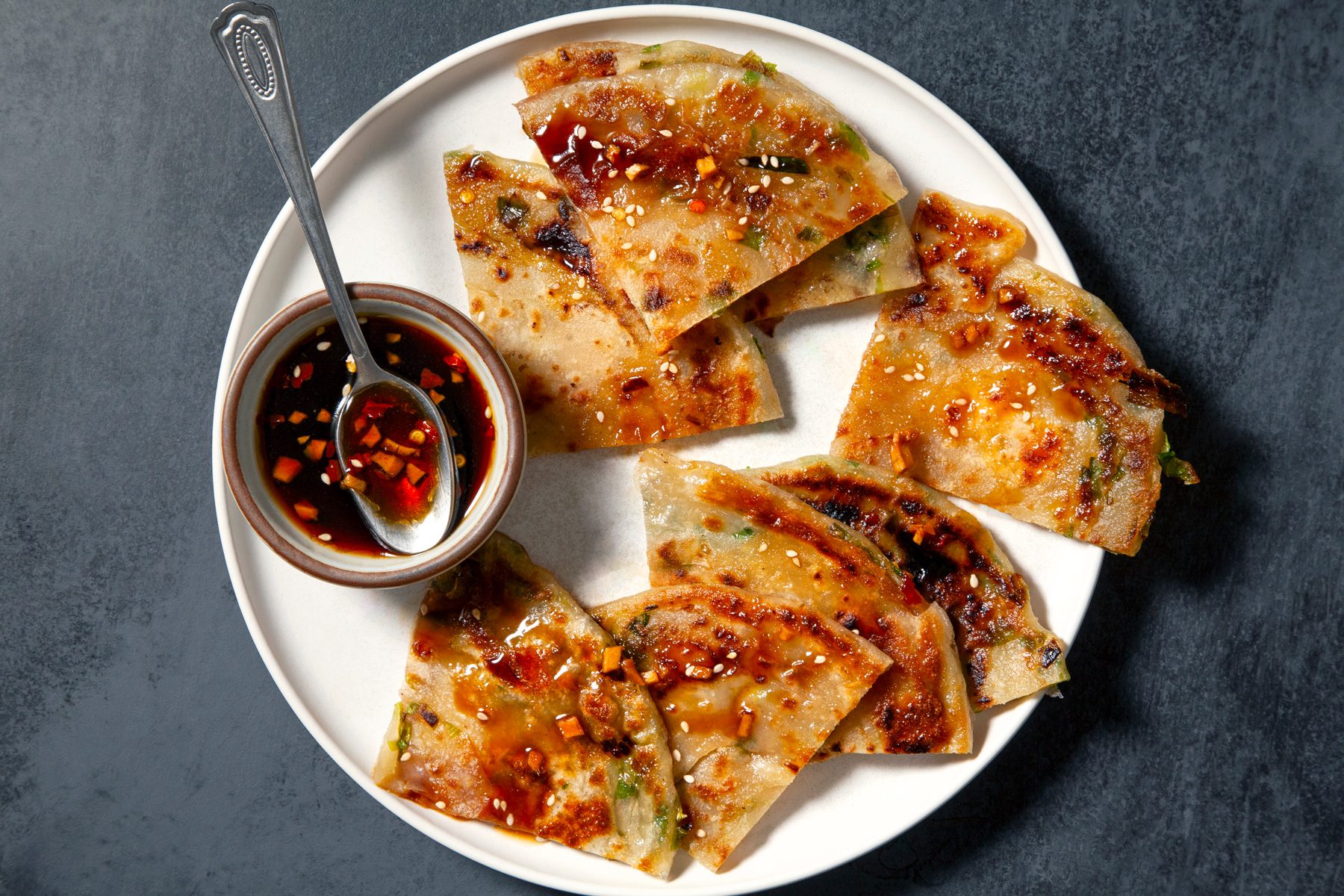 Overhead shot of Chinese scallion pancake pieces on a white plate, served with a small bowl of dipping sauce and a silver spoon, set against a grey background.