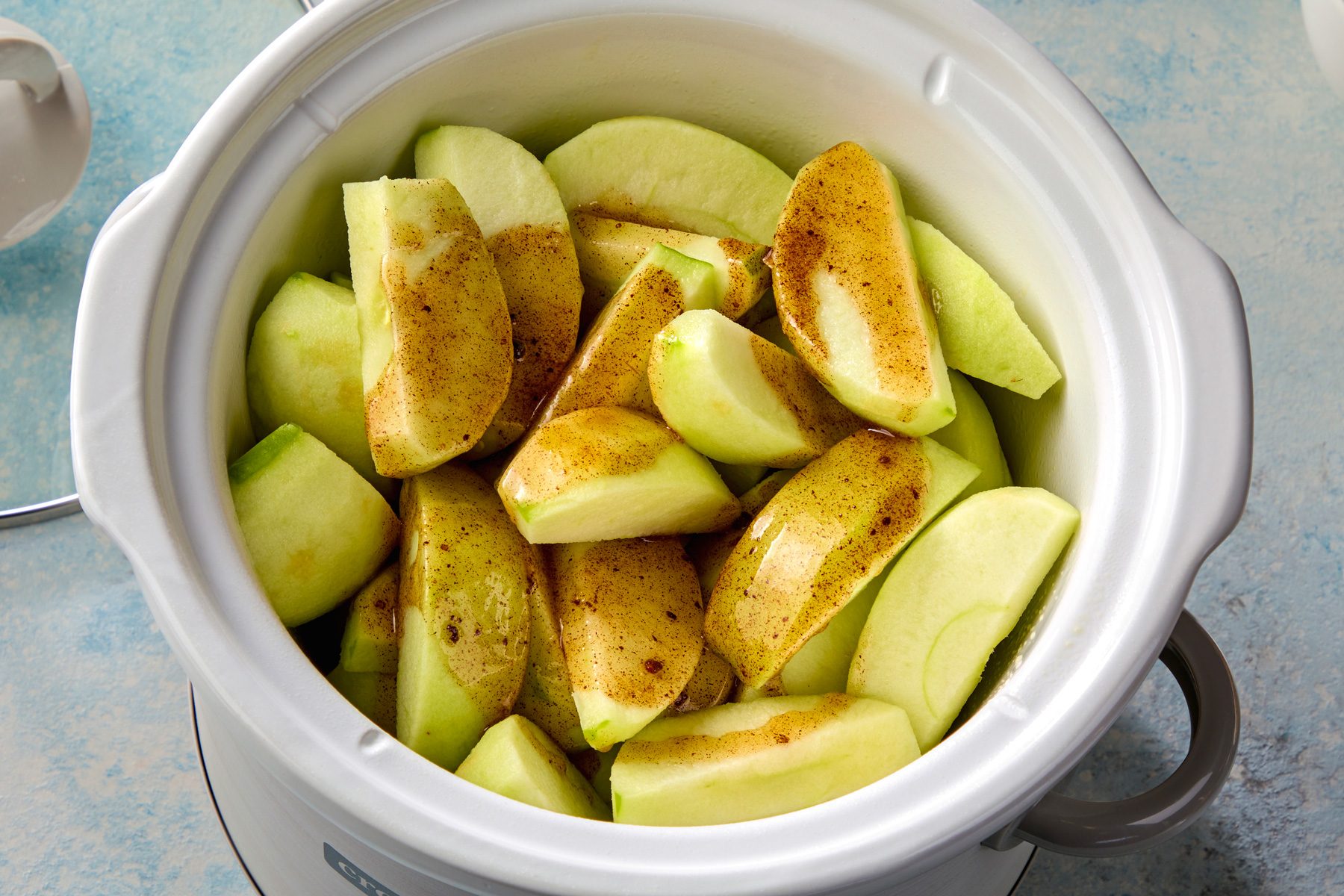 overhead shot of a white slow cooker filled with apple slices coated in a cinnamon and butter mixture; the apple slices are arranged in a single layer, with some overlapping; the slow cooker is sitting on a light blue surface;