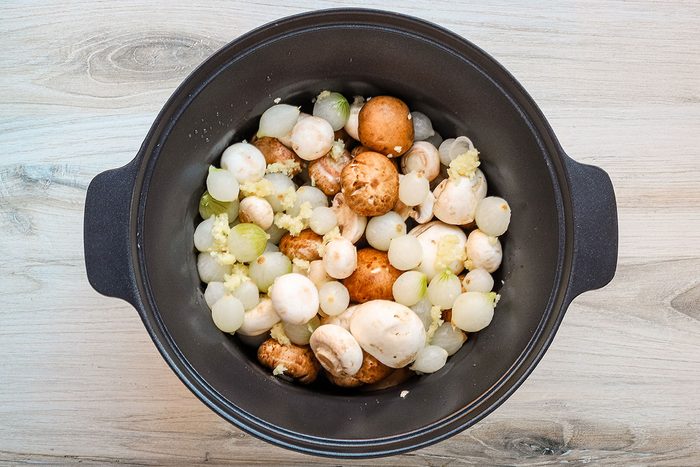 The slow cooker filled with mushrooms, pearl onions, garlic and herbs.