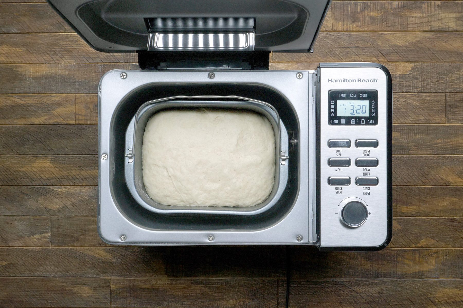 A bread machine with a batch of dough inside, displaying a digital screen reading "2:30". The machine is placed on a wooden surface, and various buttons and a knob are visible on the control panel.