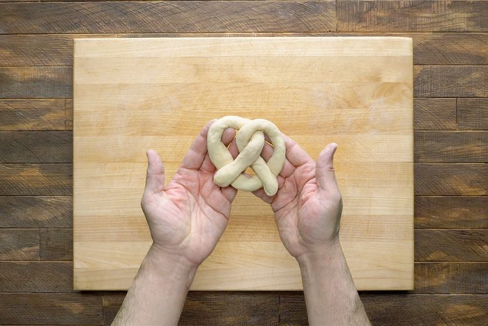 Two hands holding a raw pretzel dough in the shape of a twist above a wooden cutting board on a wooden surface.