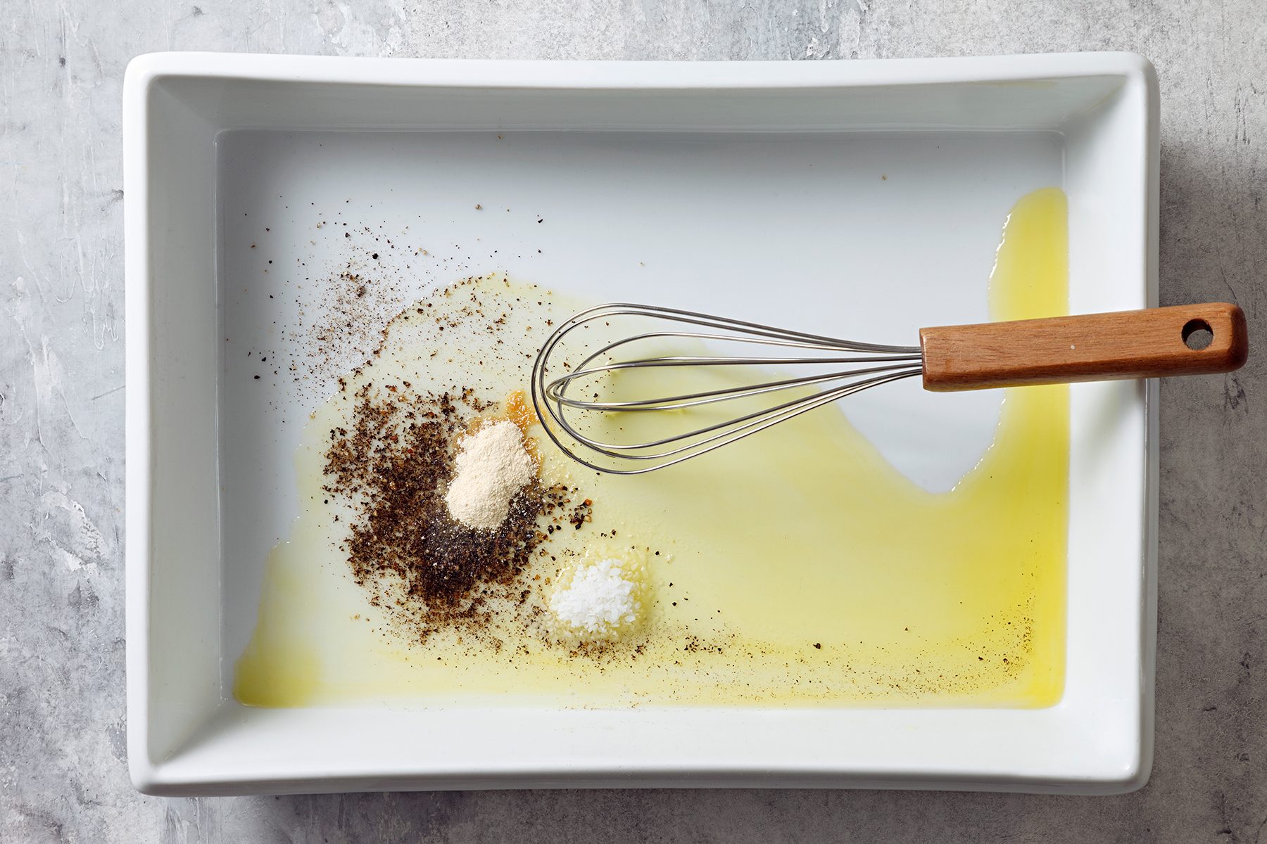 A white rectangular dish contains oil, with a whisk laying in it. Several seasonings are sprinkled next to the whisk, including a light powder and black specks, set against a neutral background.
