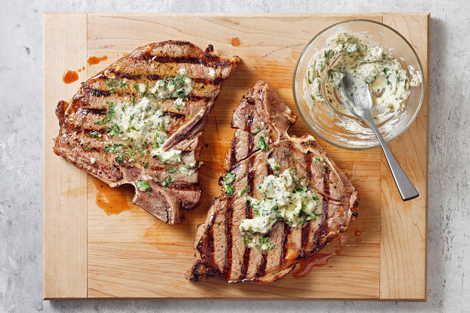 Two grilled T-bone steaks topped with herb butter are on a wooden cutting board. A glass bowl containing more herb butter and a spoon sits beside the steaks. The board rests on a light gray surface.
