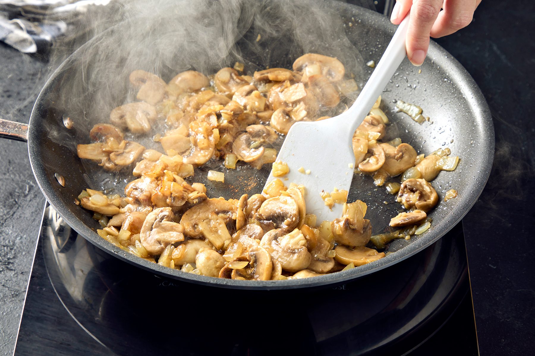 A person uses a spatula to sauté sliced mushrooms and onions in a nonstick frying pan on a stovetop. Steam rises from the pan, indicating the food is being cooked.