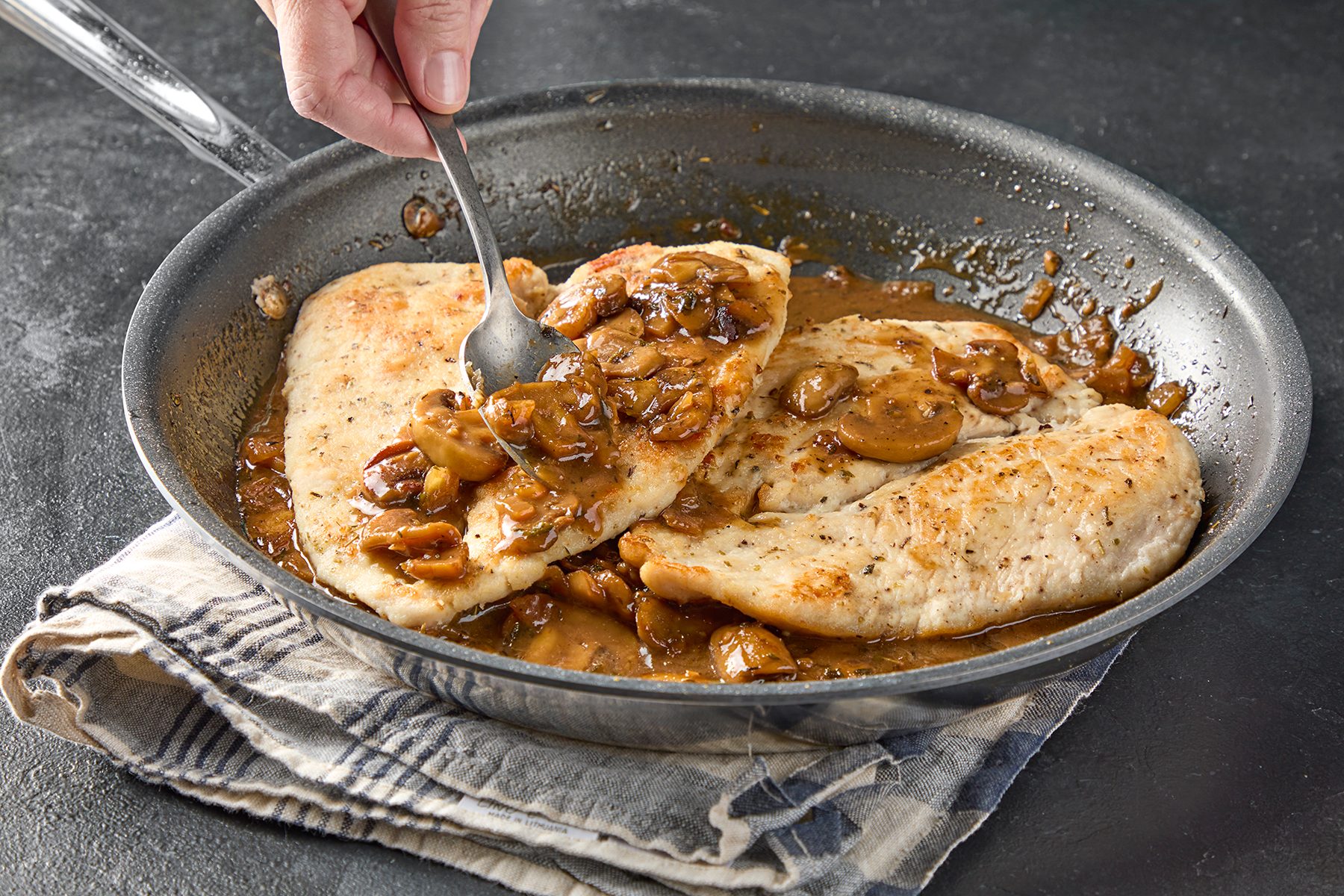 A person uses a spoon to serve chicken breasts cooked with mushrooms and sauce in a skillet. The pan rests on a striped cloth on a dark surface. .
