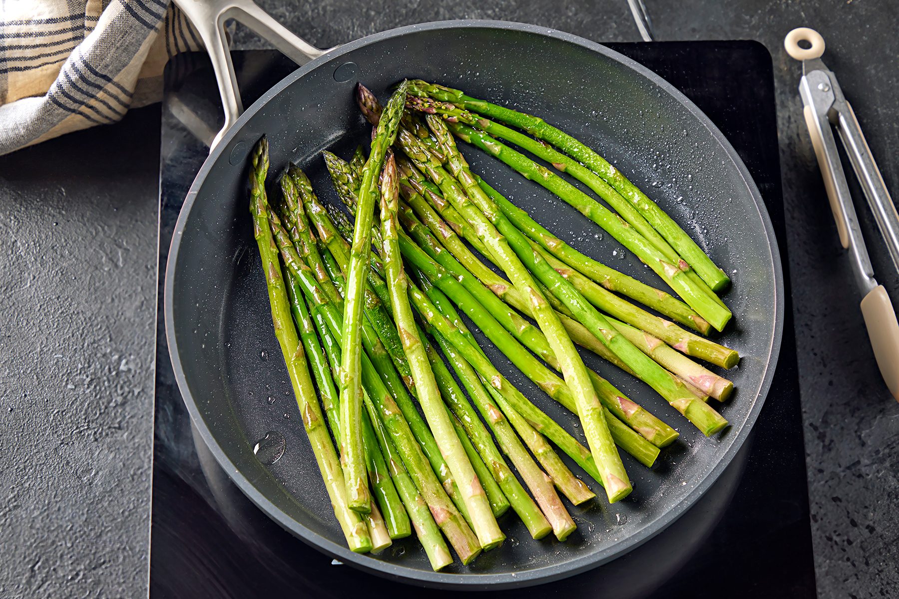 A frying pan filled with fresh asparagus spears cooking on a stovetop. The asparagus appears seasoned, and a striped kitchen towel and tongs are visible in the background on a dark countertop.