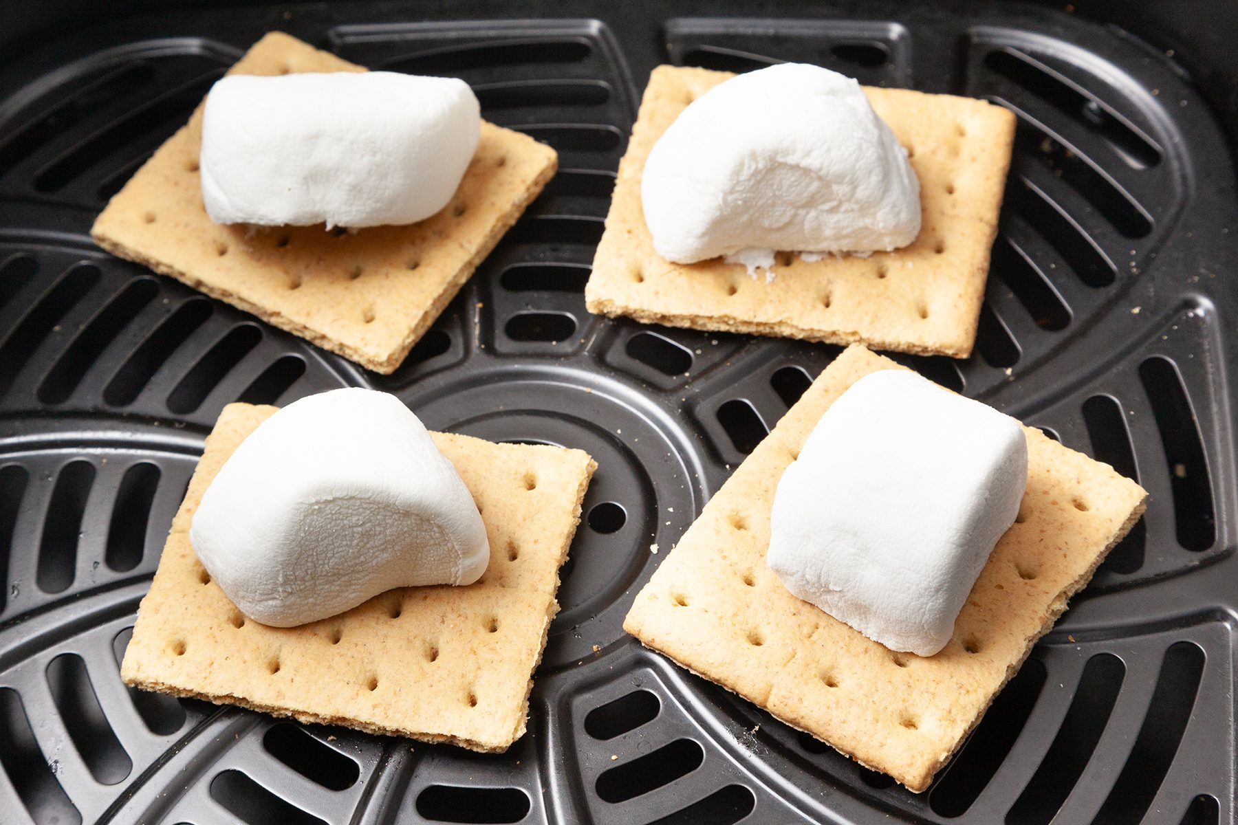 Four graham crackers topped with marshmallows are placed on an air fryer tray.