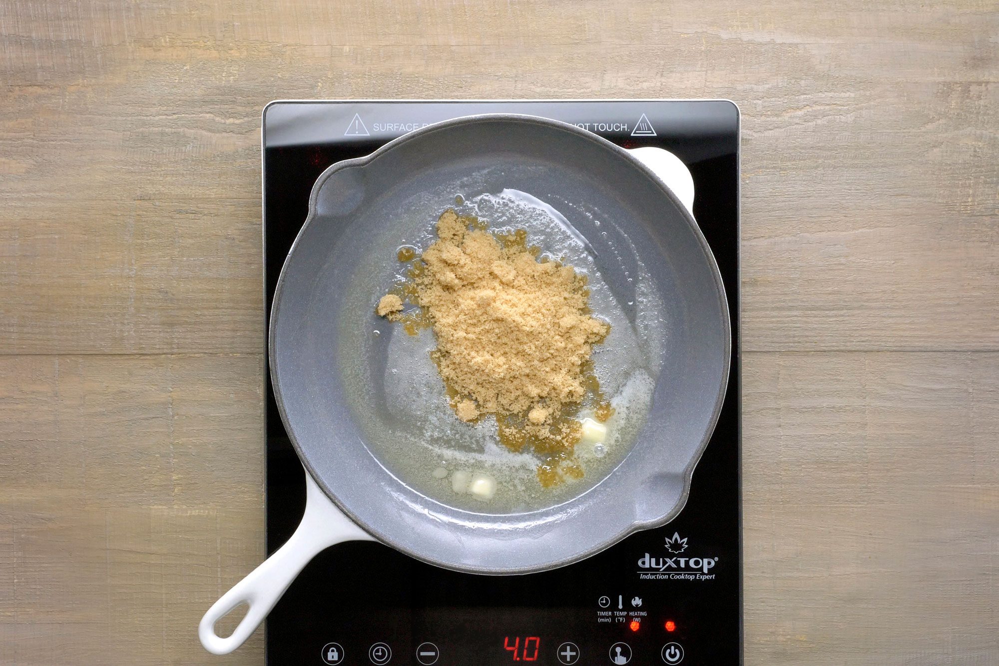 Gray cast-iron skillet sits on a black electric stovetop; Inside the skillet, a mixture of butter and brown sugar is melting together, creating a bubbling and caramelizing mixture