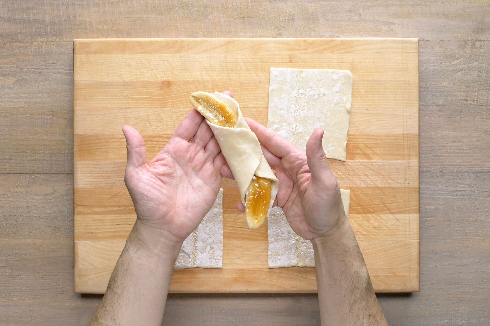 A puff pastry with a filling in it; the pastry is being rolled up with the filling inside; there are several pieces of puff pastry on a wooden cutting board next to the person's hand