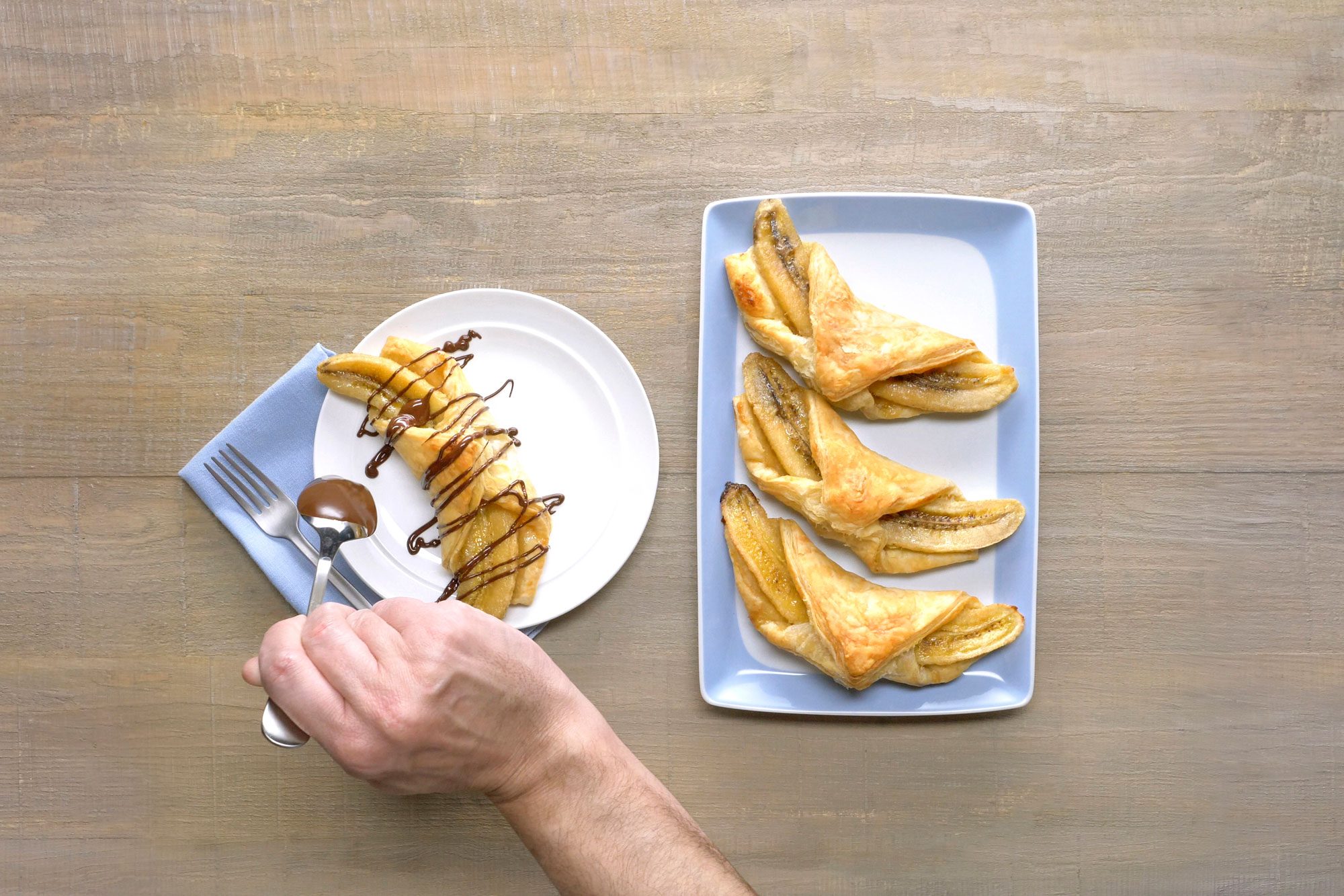 Hand is holding a spoon over a white plate with a banana pastry on it that has a chocolate sauce being drizzled over it; to the right is a blue plate with three more banana pastries on it, all in a light wooden background