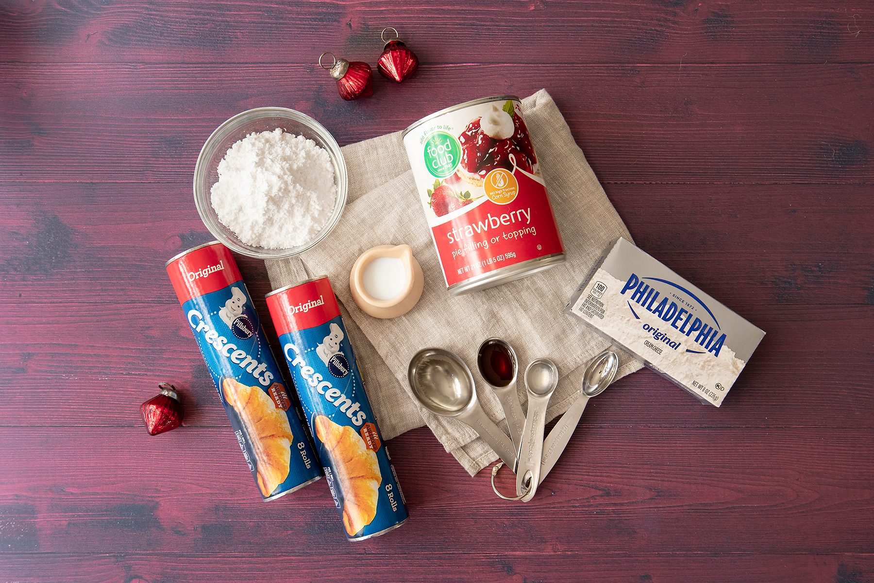 Baking ingredients on a table: Pillsbury Crescent rolls, strawberry pie filling, a block of cream cheese, a bowl of powdered sugar, a small bowl of salt, measuring spoons, a spoon, and red ornaments on a wooden surface.